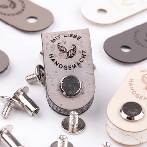 Könnte beinhalten: Nahaufnahme eines hellgrauen Lederbandes mit dem Text "Mit Liebe Handgemacht" in brauner Schrift. Das Band hat ein kleines Loch in der Mitte und ist von silbernen Metallnieten umgeben. Mehrere silberne Metallnieten sind um das Band herum verstreut.