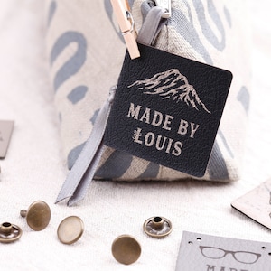 Può includere: Un'etichetta in pelle nera con un design a forma di montagna e il testo "MADE BY LOUIS" in lettere dorate. L'etichetta è attaccata a una borsa di tessuto grigio e bianco con una cerniera.