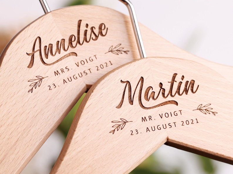 Könnte beinhalten: Zwei Holzbügel mit den eingravierten Namen "Annelise" und "Martin". Die Bügel enthalten auch den Text "MRS. VOIGT 23. AUGUST 2021" und "MR. VOIGT 23. AUGUST 2021".