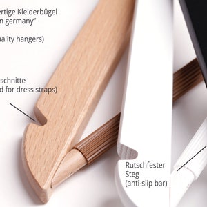 May include: Close-up of three wooden hangers with different finishes. The hanger on the left is natural wood with a notch for dress straps. The middle hanger is white with a brown anti-slip bar. The hanger on the right is black. The text "Hochwertige Kleiderb&uuml;gel "made in germany" (high quality hangers)" is visible at the top of the image. The text "Rockeinschnitte (notched for dress straps)" is visible on the left side of the image. The text "Rutschfester Steg (anti-slip bar)" is visible on the right side of the image.