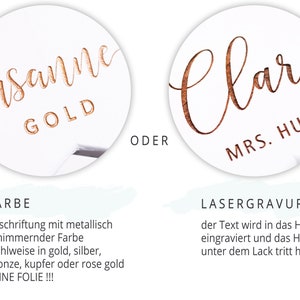 May include: Two examples of personalized wooden signs. The first sign is a white circle with the name "Susanne" in gold lettering and the word "GOLD" below. The second sign is a white circle with the name "Clara" in brown lettering and the text "MRS. HUBER" below. The text "FARBE" and "LASERGRAVUR" are written in black on a light blue background.