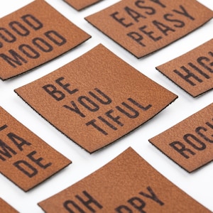Könnte beinhalten: Ein Satz von neun braunen Leder-Patches mit verschiedenen positiven Botschaften in Schwarz gedruckt. Die Botschaften beinhalten "Good Mood", "Be Youtiful", "Easy Peasy", "High 5", "Rock It", "Oh Happy Day", "Me Ma De" und mehr.