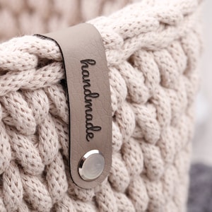Könnte beinhalten: Ein beigefarbener Häkelkorb mit einem grauen Lederanhänger, auf dem "handmade" steht.