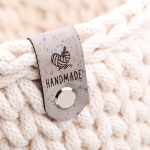 Könnte beinhalten: Nahaufnahme eines cremefarbenen, grob gestrickten Korbs. Ein graues Etikett mit dem Wort "HANDMADE" und einem herzförmigen Logo mit Garn und Nadel ist mit einem silbernen Knopf befestigt. Der Korb besteht aus dickem, gewebtem Garn.