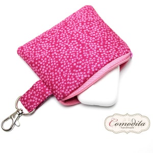 Könnte beinhalten: Eine rosa Reißverschlusstasche mit einem weißen Blumenmuster und einem silbernen Schlüsselanhänger. Die Tasche ist offen und ein weißes rechteckiges Objekt befindet sich im Inneren. Die Tasche trägt den Text "Comodita - handmade" unten.