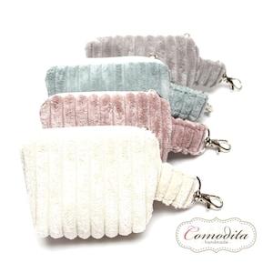 Könnte beinhalten: Fünf Reißverschlusstaschen in verschiedenen Farben, darunter beige, rosa, blau und grau. Die Taschen sind aus einem weichen, strukturierten Stoff gefertigt und haben einen silbernen Reißverschluss. Jede Tasche hat eine kleine Schlaufe zum Anbringen eines Schlüsselrings. Die Taschen sind in einer Reihe angeordnet, wobei die beige Tasche unten und die graue Tasche oben ist. Die Taschen sind alle mit den Reißverschlüssen nach rechts ausgerichtet. Die Taschen werden alle von der Firma "Comodita handmade" hergestellt.