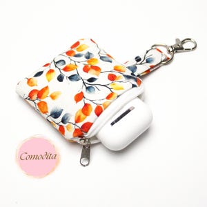 Könnte beinhalten: Weißes Stoff-AirPod-Etui mit einem bunten Blattmuster in Orange, Gelb und Blau. Das Etui hat einen Reißverschluss und einen silbernen Verschluss. Ein weißes AirPod-Etui ist teilweise sichtbar. Ein rosa Aufkleber mit dem Wort "Comodita" befindet sich unten links.