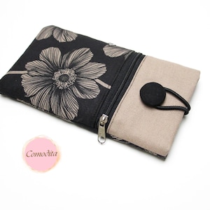 Könnte beinhalten: Eine schwarze und beige Geldbörse mit floralem Muster, Reißverschluss und Knopfverschluss. Die Geldbörse hat ein großes beiges Blumendesign auf schwarzem Hintergrund. Die andere Seite ist beige. Das Wort "Comodita" steht auf einem rosa Kreis.