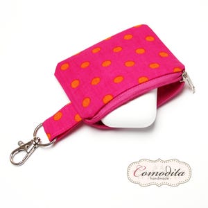 Könnte beinhalten: Eine rosa Reißverschlusstasche mit orangefarbenen Punkten und einem silbernen Schlüsselanhänger. Die Tasche ist offen und ein weißes Objekt befindet sich im Inneren.