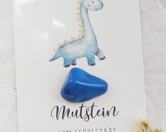 Könnte beinhalten: Eine Grußkarte mit einer Illustration eines blauen Dinosauriers und einem blauen Edelstein. Auf der Karte steht der Text "Mutstein ZUM SCHULSTART". Ein kleiner Strauß Trockenblumen befindet sich unten rechts.