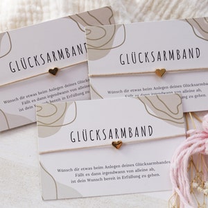 Könnte beinhalten: Eine weiße Karte mit dem Text "GLÜCKSARMBAND" und einer Botschaft darüber, dass ein Wunsch in Erfüllung geht, wenn das Armband herunterfällt. Die Karte liegt auf einer weißen Oberfläche mit einem rosa Garn-Quast.