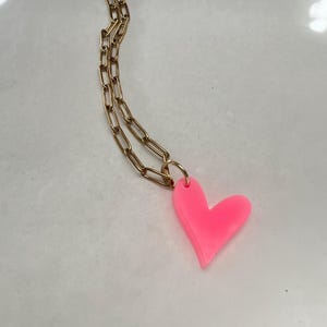 Könnte beinhalten: Eine goldfarbene Halskette mit einem leuchtend pinkfarbenen Herzanhänger. Das Herz hat eine glatte, solide Form, und die Kette hat ein Kettendesign. Die Halskette wird auf einer weißen Oberfläche präsentiert.