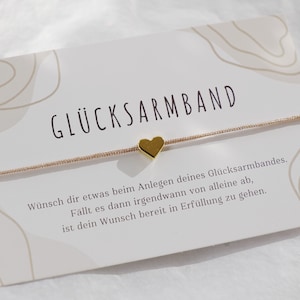 May include: A thin beige string bracelet featuring a small gold-coloured heart charm, presented on a white card with the text GLÜCKSARMBAND and German script. The card includes abstract line art in the corners and a beige colour palette.