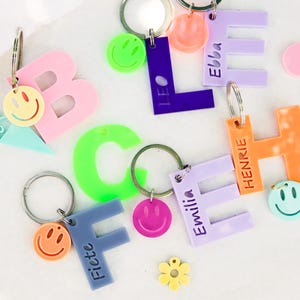 Könnte beinhalten: Eine Sammlung bunter Acryl-Buchstaben-Schlüsselanhänger in verschiedenen Farben wie Pink, Lila, Orange, Blau und Neongrün. Jeder Schlüsselanhänger hat einen Metallring und einen kleinen Smiley-Anhänger. Einige Buchstaben sind mit Namen wie Ebba, Henrie, Emilia und Fiete graviert.