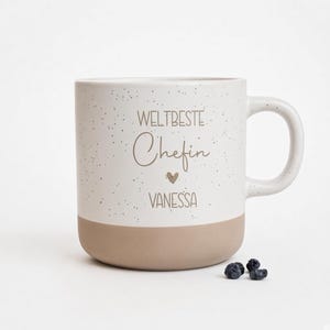 Könnte beinhalten: Ein Keramikbecher mit gesprenkeltem weißem Oberteil und einem beigen Boden. Der Text "WELTBESTE Chefin" ist in Braun aufgedruckt, gefolgt von einem kleinen Herz und dem Namen "VANESSA" in Großbuchstaben. Ein Henkel befindet sich rechts.