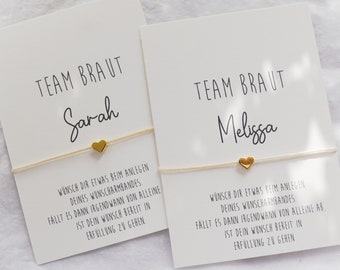 Op de afbeelding: Twee witte rechthoekige kaarten met de tekst TEAM BRAUT, de namen Sarah en Melissa en een Duits gedicht. Elke kaart bevat een dun beige koordarmbandje met een klein, gepolijst goudkleurig metalen hartje in het midden van de kaart.