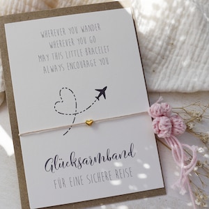 Puede incluir: Una tarjeta blanca con el texto "Wherever you wander wherever you go may this little bracelet always encourage you" y "Glucksarmband fur eine sichere Reise". Una pulsera de cuerda blanca con un dije de corazón dorado y un pequeño dije de avión está unida a la tarjeta.