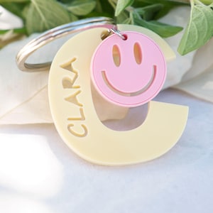 Puede incluir: Un llavero de acrílico color crema con forma de letra C y el nombre CLARA grabado verticalmente. Un pequeño dije circular de color rosa claro con una cara sonriente recortada está sujeto a la anilla metálica junto a la letra C.
