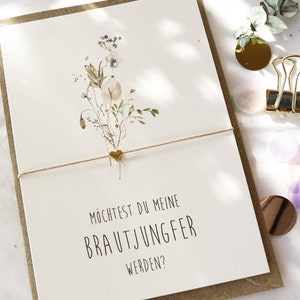 Könnte beinhalten: Eine Grußkarte mit floralem Design und dem Text "Möchtest du meine Brautjungfer werden?" in Schwarz. Ein goldener Herzanhänger ist mit einer Schnur an der Karte befestigt. Die Karte befindet sich auf einem braunen Umschlag.