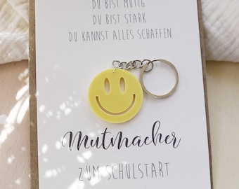 Könnte beinhalten: Ein gelber Smiley-Schlüsselanhänger mit dem Text "Du bist mutig, Du bist stark, Du kannst alles schaffen" und "Mutmacher zum Schulstart" auf einer weißen Karte.