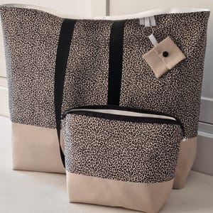 Könnte beinhalten: Ein großes Shopper-Set und eine passende Kosmetiktasche. Die Taschen haben ein schwarz-braunes Leopardenmuster mit schwarzen Trägern und einem beigefarbenen Boden. Ein kleines, beigefarbenes Täschchen mit schwarzem Knopf ist an der Tragetasche befestigt.
