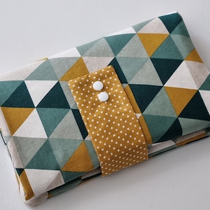 May include: A folded fabric changing mat with a geometric pattern of green, white, and mustard yellow triangles. The mat has a mustard yellow flap with white polka dots and two white buttons.
