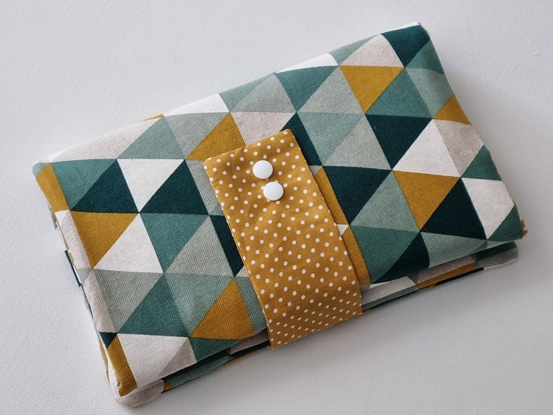 May include: A folded fabric changing mat with a geometric pattern of green, white, and mustard yellow triangles. The mat has a mustard yellow flap with white polka dots and two white buttons.