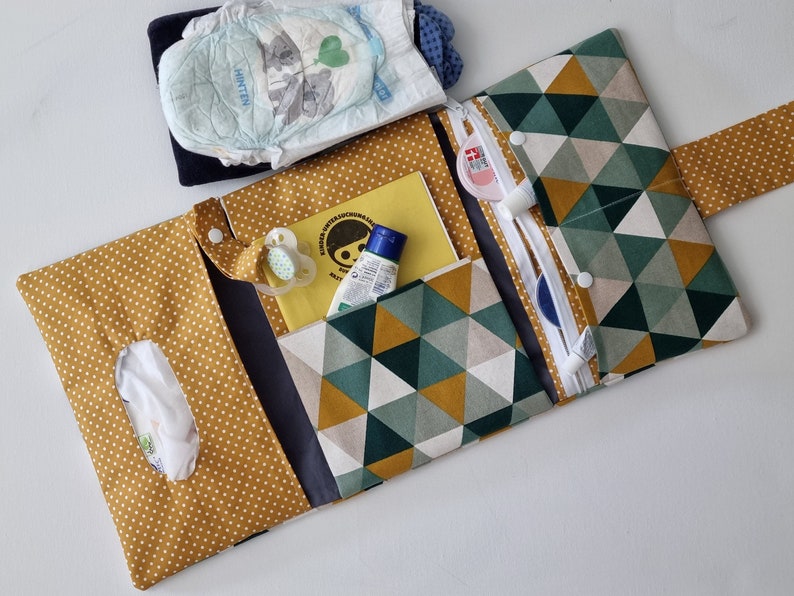 May include: A yellow and green patterned diaper bag with multiple pockets and compartments. The bag is open and contains a diaper, wipes, a pacifier, lotion, and a small book. The bag has a zipper closure and a strap.