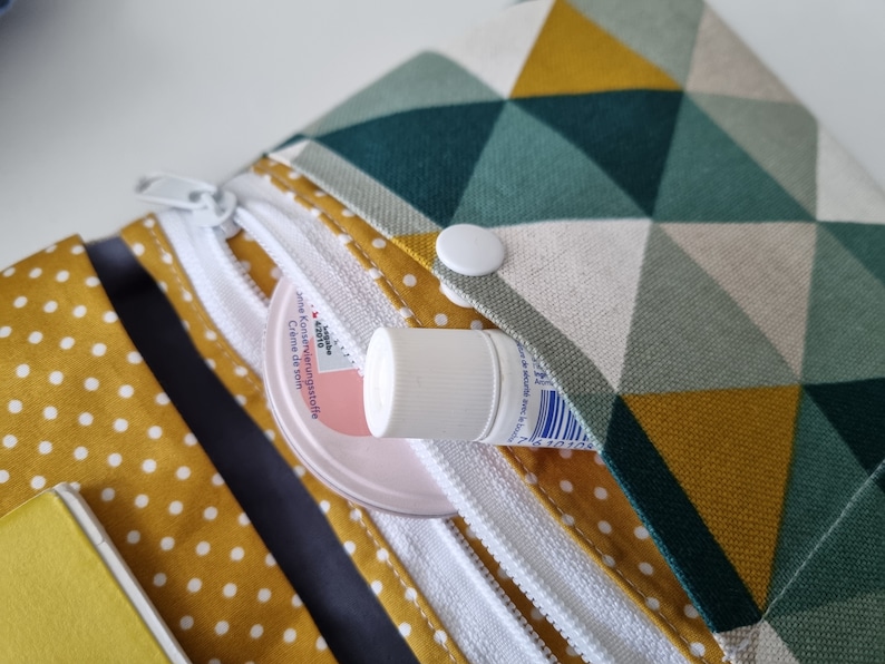 May include: A small, white zippered pouch with a geometric pattern in shades of green, yellow, and white. The pouch has a white button closure and a small tube of cream is partially visible inside.