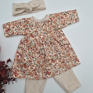 Peut inclure: Un ensemble de vêtements pour poupée bébé avec une robe à imprimé floral, un legging beige et un bandeau beige.