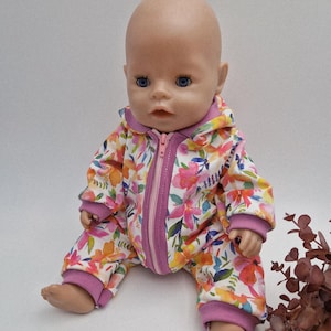 Könnte beinhalten: Eine Babypuppe in einem floral bedruckten Overall mit Kapuze und Reißverschluss. Der Overall hat ein Muster aus rosa, orangefarbenen und gelben Blumen mit grünen Blättern auf weißem Grund. Die Bündchen und der Besatz sind einfarbig rosa.