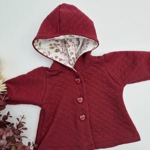 Puede incluir: Una chaqueta con capucha color burdeos con un patrón acolchado y botones en forma de corazón. La capucha está forrada con una tela blanca con estampado floral.
