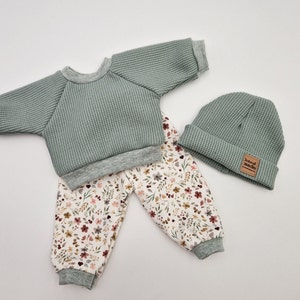 Op de afbeelding: Een driedelige babyoutfit in saliegroen en bloemenprint. De set bevat een top met lange mouwen van wafelstof, een muts met een "hand made" label en een broek met bloemenprint en grijze manchetten. De outfit is voor baby's.