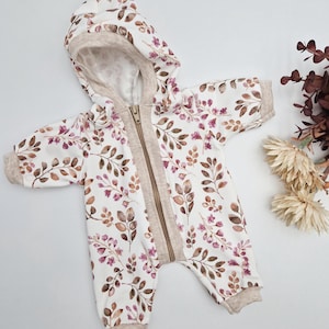 Könnte beinhalten: Weißer Baby-Overall mit floralem Muster in Braun- und Lilatönen. Der Kapuzen-Overall hat einen durchgehenden Reißverschluss, beige Verzierungen und gerippte Bündchen. Das Kleidungsstück ist für Säuglinge und Kleinkinder konzipiert.