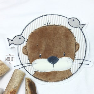 Könnte beinhalten: Ein weißes T-Shirt mit einem gestickten Otter-Design. Der Otter ist braun mit einer weißen Schnauze und ist von einem gestreiften Kreis mit zwei grauen Fischen umgeben. Der Text "MEBRIT" befindet sich darunter.