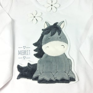 Könnte beinhalten: Weißes Langarmshirt mit einem grauen Pferde-Applikation. Das Pferd hat eine dunkelgraue Mähne und einen Schweif, darunter steht der Name "MEBRIT". Weiße Blumenakzente befinden sich am Ausschnitt.