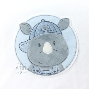 Könnte beinhalten: Ein kuscheliges, graues Nashorn mit einer blauen Jeans-Baseballkappe und Latzhose, vor einem hellblauen kreisförmigen Hintergrund. Das Nashorn hat eine weiße Nase und lächelt. Das Bild hat den Text "MEBRIT" in der linken unteren Ecke.