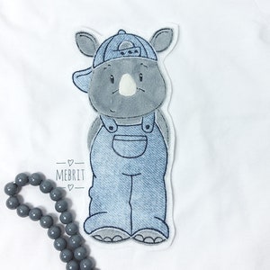 Könnte beinhalten: Ein graues Nashorn mit einer blauen Jeansmütze und Latzhose. Das Nashorn hat ein weißes Horn und lächelt. Das Bild enthält den Text "MEBRIT" und eine Kette aus grauen Perlen.
