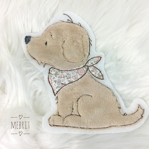 Könnte beinhalten: Ein hellbraunes, plüschiges Hundekissen mit einem floral gemusterten Halstuch. Der Hund hat eine schwarze Nase und gestickte Details. Das Kissen befindet sich auf weißem Hintergrund. Der Markenname "MEBRIT" steht unten.