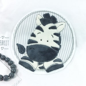 Könnte beinhalten: Runder Aufnäher mit einem lächelnden Zebra-Design. Das Zebra ist in Schwarz und Weiß gehalten und befindet sich vor einem grau-weiß gestreiften Hintergrund. Das Wort "MEBRIT" ist im Bild enthalten.