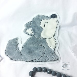Könnte beinhalten: Ein grau-weißes Wolf-Applikation ist auf ein weißes T-Shirt genäht. Der Wolf sitzt mit zurückgeneigtem Kopf, als würde er heulen. Die Applikation hat eine weiche, plüschige Textur. Der Markenname "MEBRIT" ist darunter gedruckt.