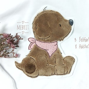 Könnte beinhalten: Ein Applikation eines braunen Plüschwelpen mit einem rosa gepunkteten Halstuch auf einer weißen Oberfläche. Die Applikation hat eine genähte Kontur und der Text "MEBRIT" befindet sich über dem Welpen. Der Text "3 Fellfarben" und "8 Halstuchfarben" sind ebenfalls sichtbar.