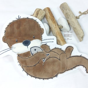 Könnte beinhalten: Ein braunes und weißes Plüschotter-Spielzeug, das einen grauen Fisch hält. Der Otter liegt auf dem Rücken, mit schwarzen Nähten. Das Bild enthält Treibholz und den Text "MEBRIT".