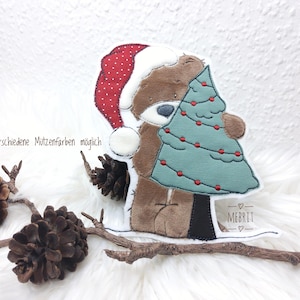 Könnte beinhalten: Ein brauner Teddybär mit Weihnachtsmütze, der einen tealfarbenen Weihnachtsbaum mit roten Ornamenten hält. Das Bild enthält den Text "verschiedene Mützenfarben möglich" und den Markennamen "MEBRIT".