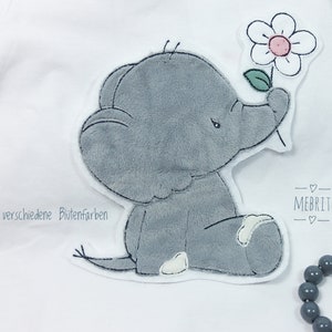 Könnte beinhalten: Ein weißer Body mit einem grauen Elefanten-Applikation, der eine Blume mit rosa Mitte und grünem Blatt hält. Der Text "8 verschiedene Blütenfarben" ist sichtbar. Ein graues Perlenarmband befindet sich im Hintergrund.