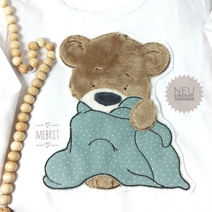 Könnte beinhalten: Weißes Langarmshirt mit einem braunen Teddybär-Applikation. Der Bär ist in eine hellblaue Decke mit weißen Punkten gehüllt. Der Text "MEBRIT" steht unter dem Bären. Eine Holzkette befindet sich links.