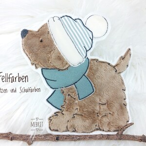 Könnte beinhalten: Ein Plüschhundespielzeug mit hellbraunem Körper, das eine blau-weiß gestreifte Mütze und einen blauen Schal trägt. Das Bild enthält den Text "3 Fellfarben" und "5 Mützen und Schalfarben". Das Spielzeug liegt auf einer weißen, flauschigen Oberfläche.