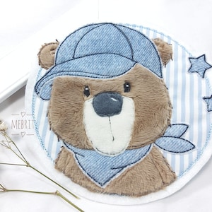 Könnte beinhalten: Gestickter Aufnäher mit einem braunen Teddybären, der eine blaue Jeansmütze und ein Halstuch trägt. Der Bär ist vor einem blau-weiß gestreiften Hintergrund mit drei blauen Sternen platziert. Der Aufnäher hat den Text "MEBRIT" unten links.