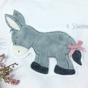 Könnte beinhalten: Weißes Langarmshirt mit einem grauen Esel-Applikation. Der Esel hat ein weißes Gesicht, einen grauen Körper und dunkelgraue Hufe. Eine rosa Schleife ist am Schwanz des Esels befestigt. Der Text "3 Schleifenfarben" steht über dem Esel.