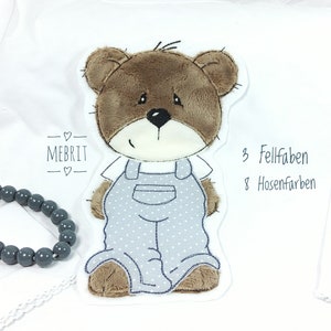 Könnte beinhalten: Ein weißes Langarmshirt mit einem braunen Teddybär-Applikation, der eine blaue Latzhose mit weißen Punkten trägt. Der Text "MEBRIT" befindet sich über dem Bären. Der Text "3 Fellfarben 8 Hosenfarben" befindet sich rechts vom Bären.